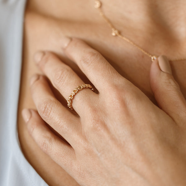 Close up of a hand with a gold floral-etched ring band with textured violet flowers and small clear gemstones, with a matching necklace being worn in the background.