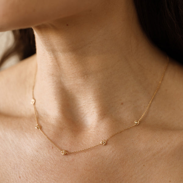 A lifestyle image of a dainty gold necklace on a person's neck with stationary textured violet flowers with small accent stones in the center along the chain.