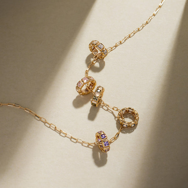 A gold paperclip link necklace with round pendants, each featuring square cut bezel-set gemstones of different colors, on a light background