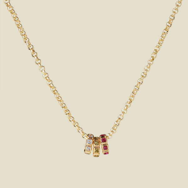 A delicate gold chain necklace with round links and three round gold ring charms attached, each with a different color gemstone, light blue, red and light green. The necklace is hanging in front of a solid light beige background.