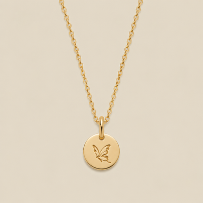 A gold chain necklace with a round gold disc pendant engraved with a butterfly, displayed against a solid beige background.