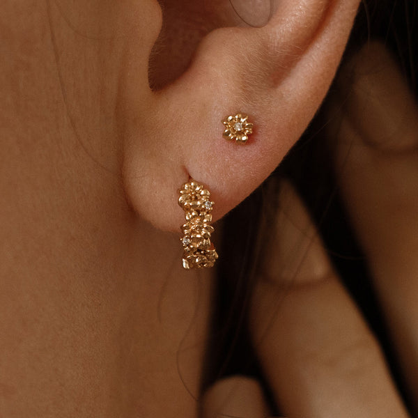 Close-up of a person wearing gold hoop earrings made up of a textured floral design and a matching stud earring with a singular flower and one accent stone in the center.