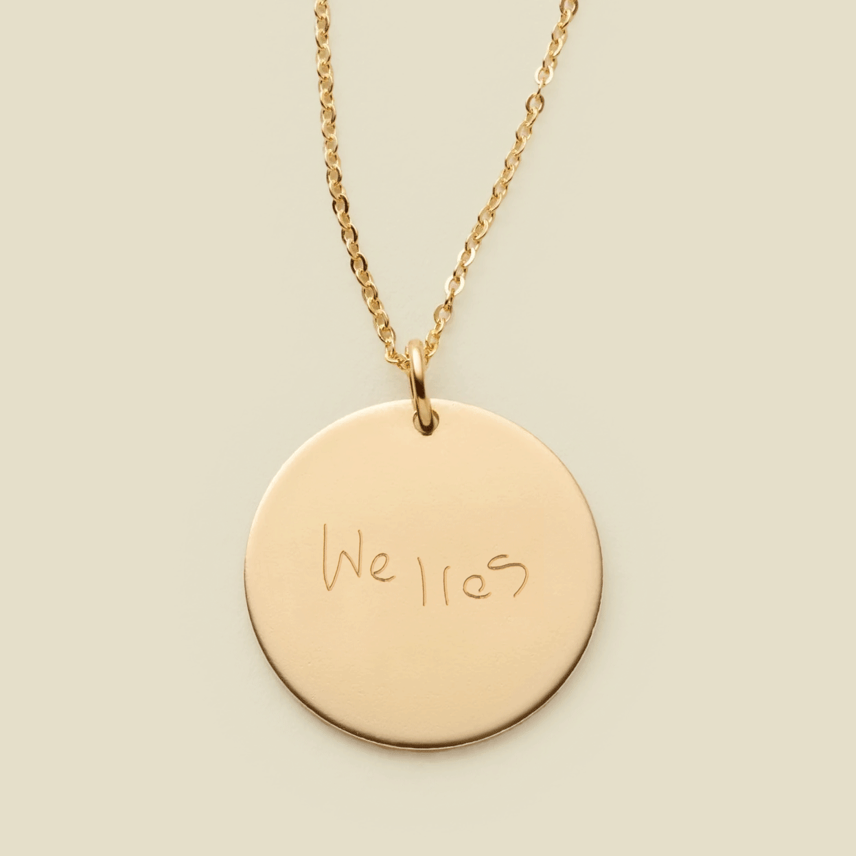 A dainty gold chain necklace with a round gold pendant engraved with handwritten script.