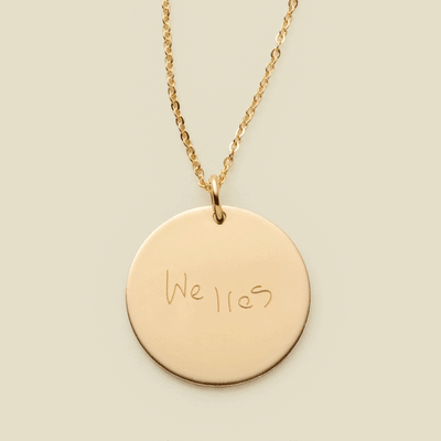 A dainty gold chain necklace with a round gold pendant engraved with handwritten script.