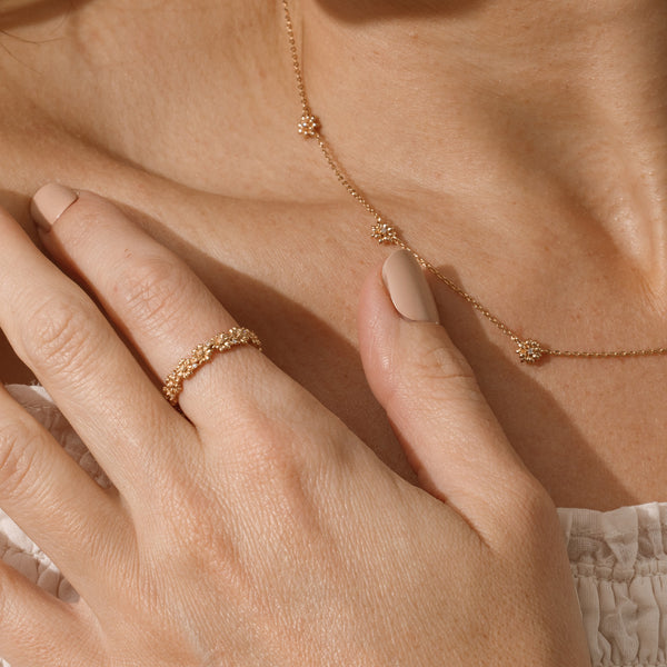 Close up of a hand with a gold floral-etched ring band with textured peony flowers and small clear gemstones, with a neck wearing the matching birth flower stationary necklace.