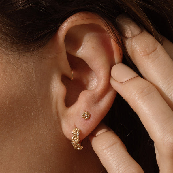Close-up of a person wearing gold hoop earrings made up of a textured floral design and a matching stud earring with a singular floral motif, with a hand adjusting them.