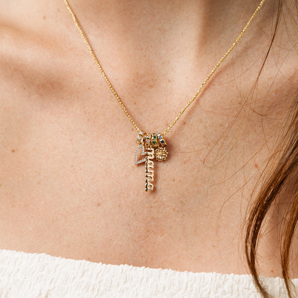 A gold chain necklace with four gold ring charms with different colored gemstones, a heart charm with clear stones, a script charm with the word 'mama', and a flower charm.
