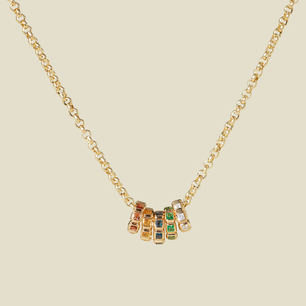 A gold vermeil necklace with a series of multicolored birthstone spacer charms, set against a solid beige background.