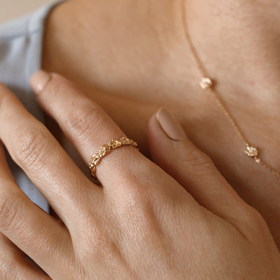 Close up of a hand with a gold floral-etched ring band with textured lily of the valley flowers and small clear gemstones, with the matching necklace being worn in the background.