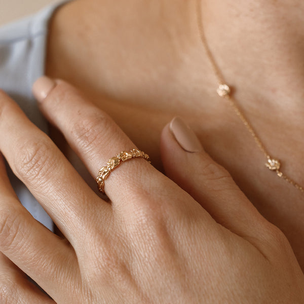 Close up of a hand with a gold floral-etched ring band with textured lily of the valley flowers and small clear gemstones, with the matching necklace being worn in the background.