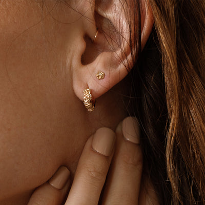 Close-up of a person wearing gold hoop earrings made up of a textured floral design and a matching stud earring with a singular floral motif.
