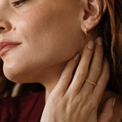 Close up of a hand with a gold floral-etched ring band with textured rose flowers and small clear gemstones, next to an ear with matching hoops and studs.