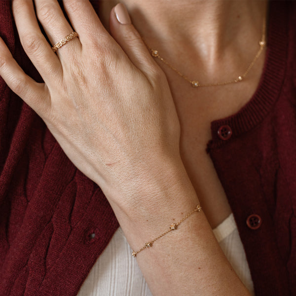A lifestyle image of a dainty gold bracelet on a person's wrist with stationary textured rose flowers with small accent stones in the center along the chain, also featuring the matching necklace and ring.