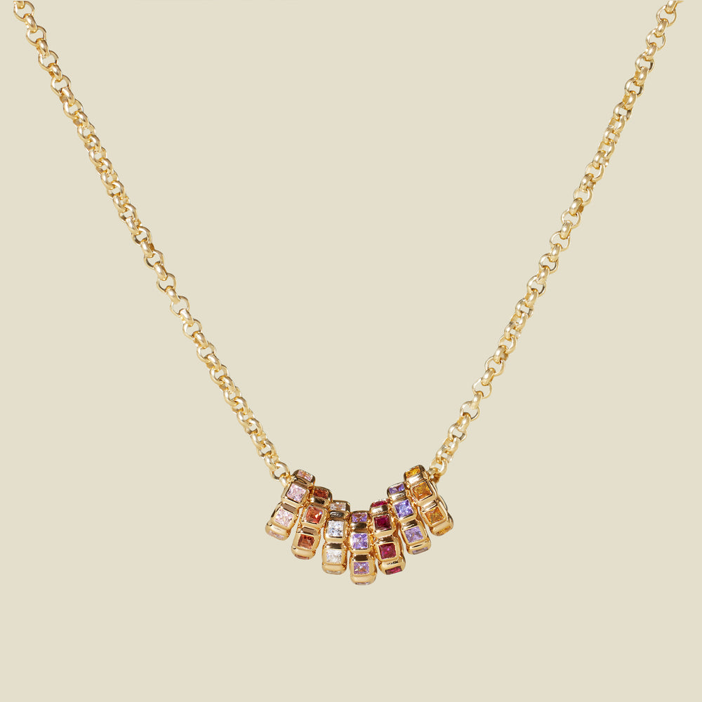 A gold vermeil necklace with a series of multicolored birthstone spacer charms, set against a solid beige background.