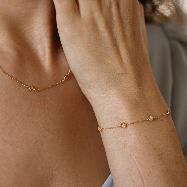 A lifestyle image of a dainty gold bracelet on a person's wrist with stationary textured poppy flowers with small accent stones in the center along the chain, also featuring the matching necklace on the model's neck.