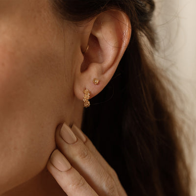A close up of a person's ear wearing a gold hoop earring made up of stacked flowers, and a matching stud with a singular flower and accent stone in the center.