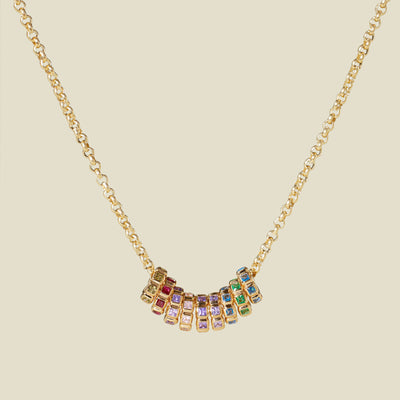 A gold vermeil necklace with a series of multicolored birthstone spacer charms, set against a solid beige background.