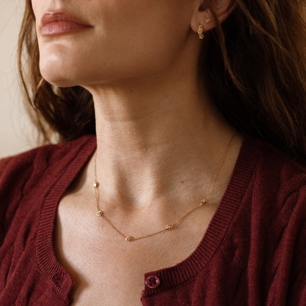A model in a red cardigan wearing a dainty gold peony station necklace with clear stones and matching floral hoop and stud earrings.