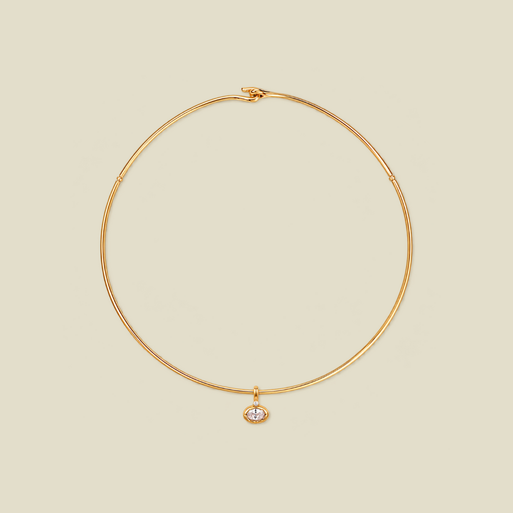A delicate gold wire circle necklace with a small oval bezel-set clear gemstone pendant, displayed on a solid light beige background.