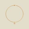 A delicate gold wire circle necklace with a small oval bezel-set clear gemstone pendant, displayed on a solid light beige background.