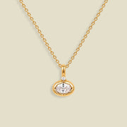 A gold chain necklace with a small oval pendant featuring a clear faceted gemstone and a tiny clear accent stone above it, shown on a light beige background.