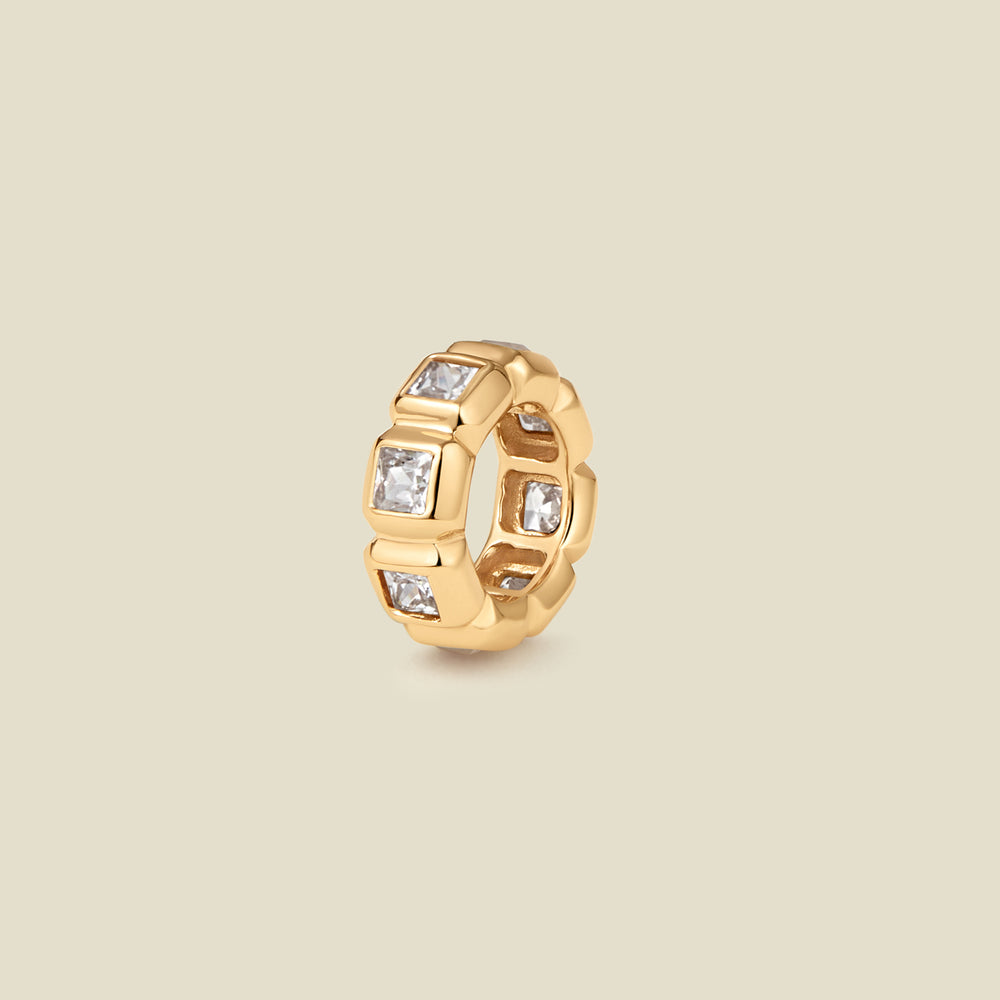 A gold ring charm with multiple bezel set square-cut clear gemstones set side by side to create the band, shown on a solid beige background.