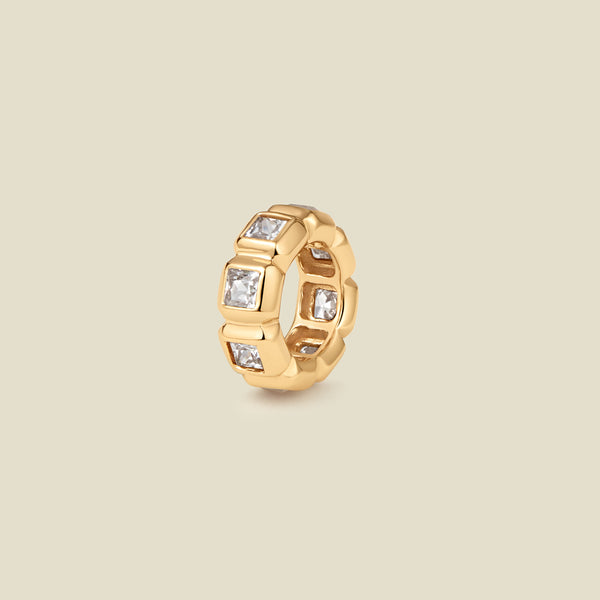 A gold ring charm with multiple bezel set square-cut clear gemstones set side by side to create the band, shown on a solid beige background.