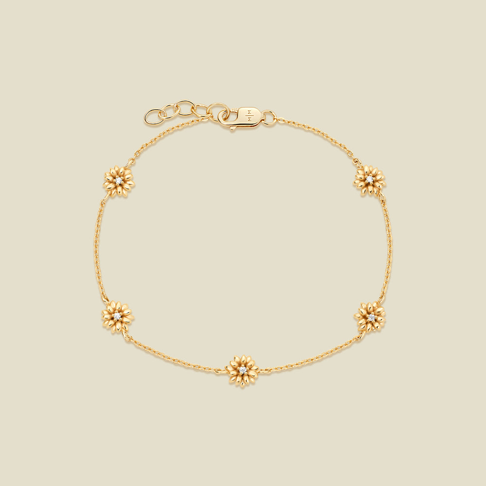 A delicate gold chain bracelet with five small daisy charms, each accented with a small clear stone in the center, displayed on a light beige background.