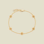 A delicate gold chain bracelet with five small daisy charms, each accented with a small clear stone in the center, displayed on a light beige background.