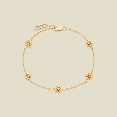 A delicate gold chain bracelet with five small daisy charms, each accented with a small clear stone in the center, displayed on a light beige background.
