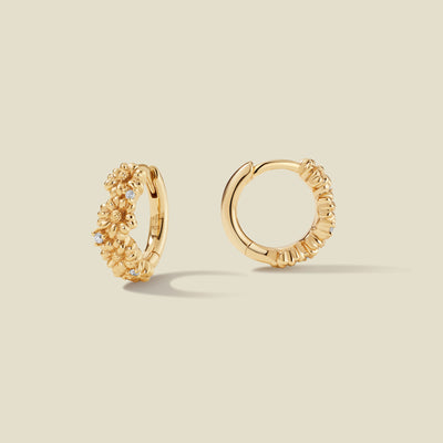 A pair of gold hoop earrings adorned with textured daisy flowers stacked along the hoop, with scattered small clear accent stones, set against a solid beige background.