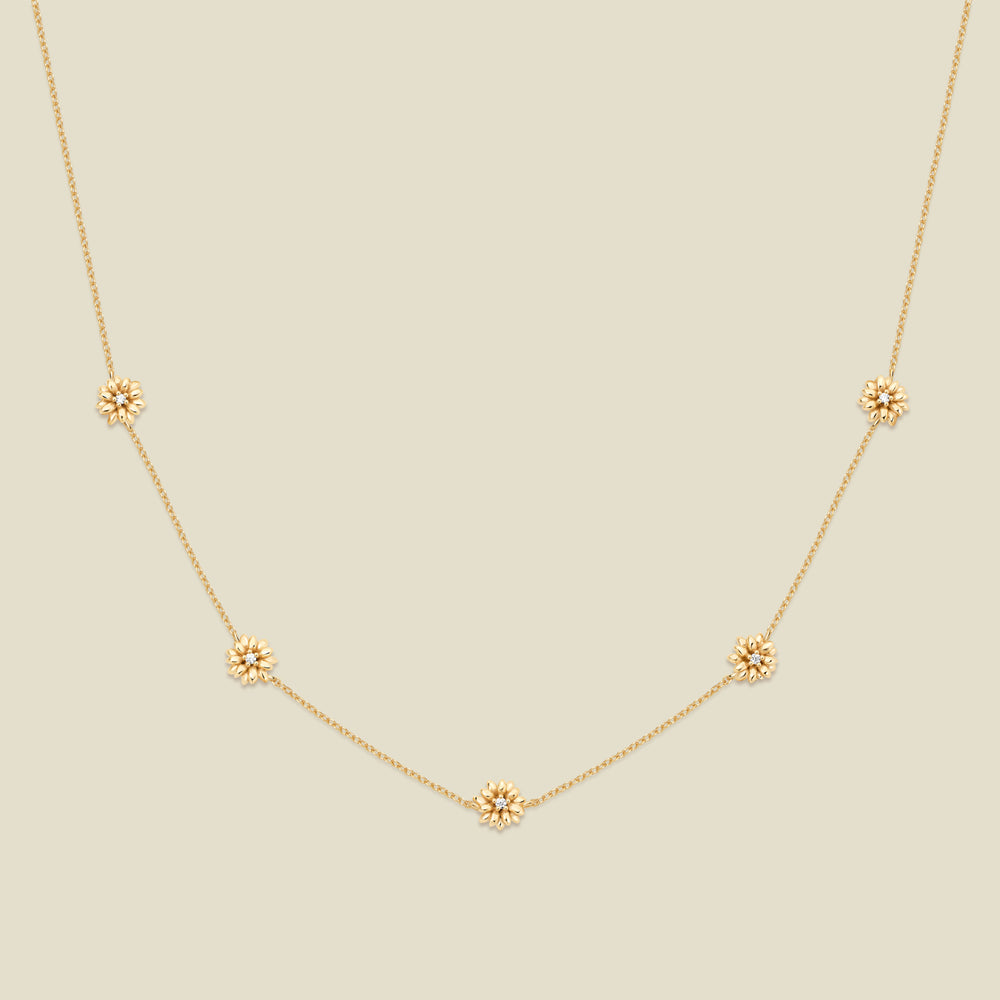 A delicate gold chain necklace featuring five small, evenly spaced textured daisy flower motifs set with tiny clear stones, displayed on a solid light beige background.