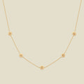 A delicate gold chain necklace featuring five small, evenly spaced textured daisy flower motifs set with tiny clear stones, displayed on a solid light beige background.