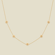 A delicate gold chain necklace featuring five small, evenly spaced textured daisy flower motifs set with tiny clear stones, displayed on a solid light beige background.