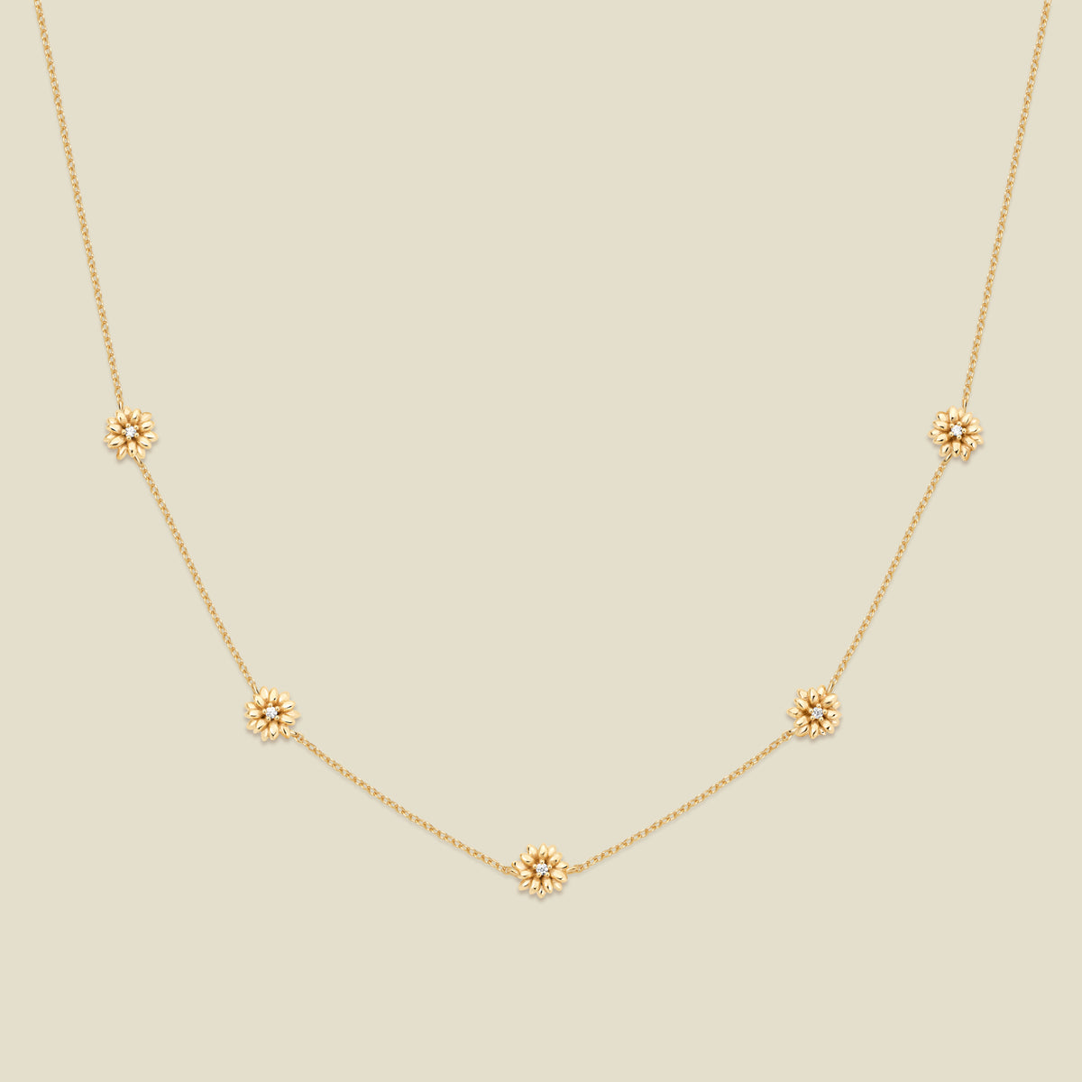 A delicate gold chain necklace featuring five small, evenly spaced textured daisy flower motifs set with tiny clear stones, displayed on a solid light beige background.