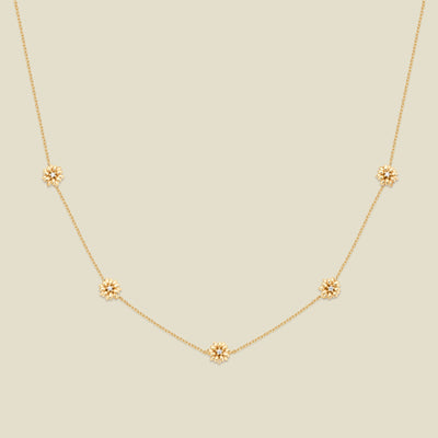 A delicate gold chain necklace featuring five small, evenly spaced textured daisy flower motifs set with tiny clear stones, displayed on a solid light beige background.