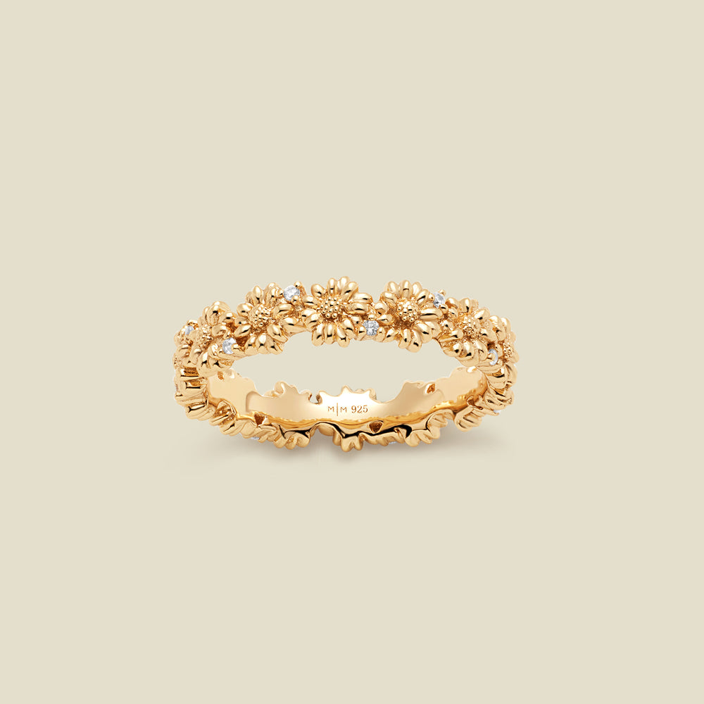 A gold floral-etched ring band with textured daisy flowers and small clear gemstones, shown on a plain warm beige background. 