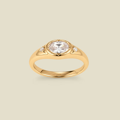 A gold signet ring with a clear center oval bezel-set gemstone, and two small clear accent stones on either side, set against a solid beige background.