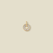 A circular gold pendant featuring clear baguette-cut stones around a hollow center, displayed against a solid light beige background.