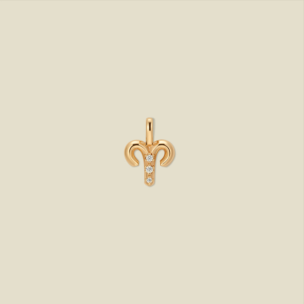 A gold pendant shaped like the Aries zodiac symbol, featuring three small clear gemstones, centered on a solid light beige background.