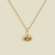 A delicate gold chain necklace with a small oval pendant featuring a faceted olive-green gemstone and a tiny clear accent stone above it, displayed on a solid light beige background.