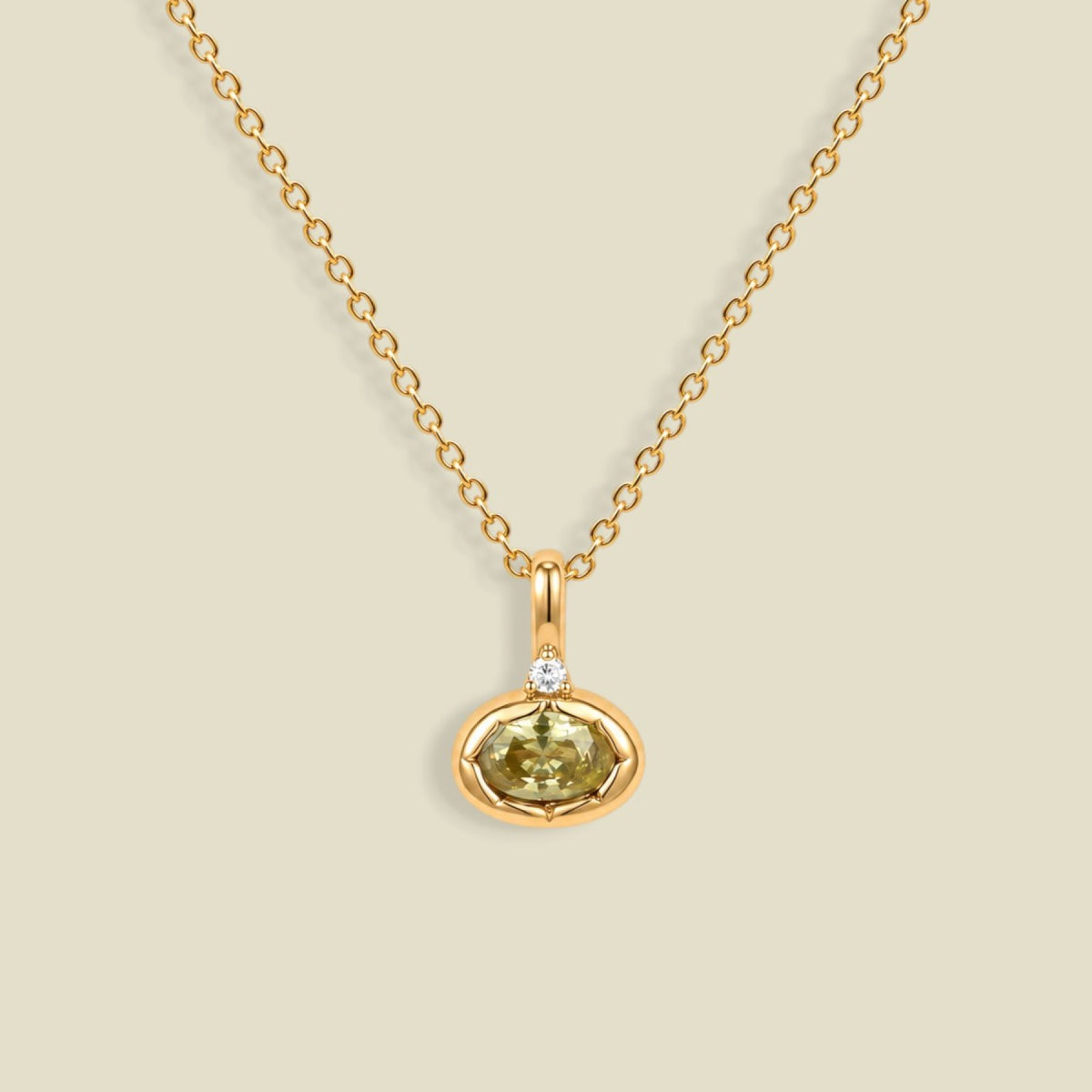A delicate gold chain necklace with a small oval pendant featuring a faceted olive-green gemstone and a tiny clear accent stone above it, displayed on a solid light beige background.