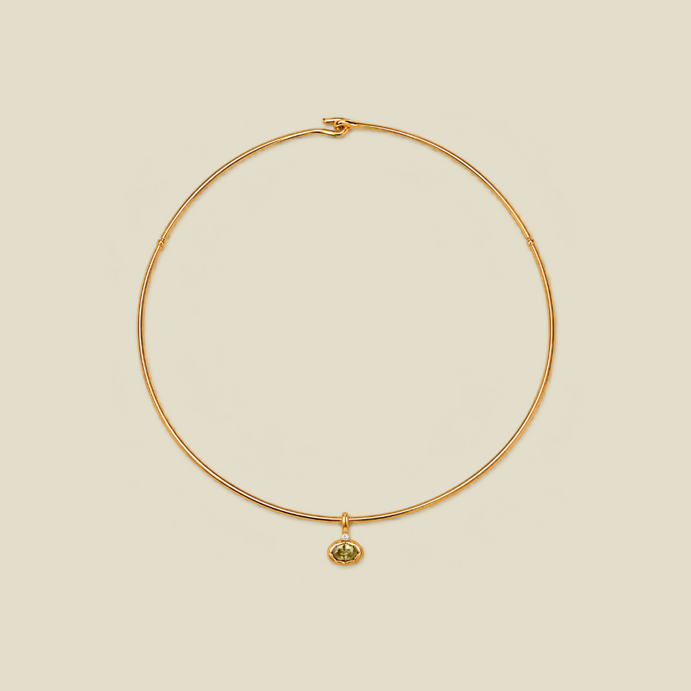 A delicate gold wire necklace with a small oval pendant featuring a green gemstone, displayed on a solid light beige background.