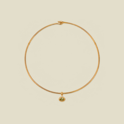 A delicate gold wire necklace with a small oval pendant featuring a green gemstone, displayed on a solid light beige background.