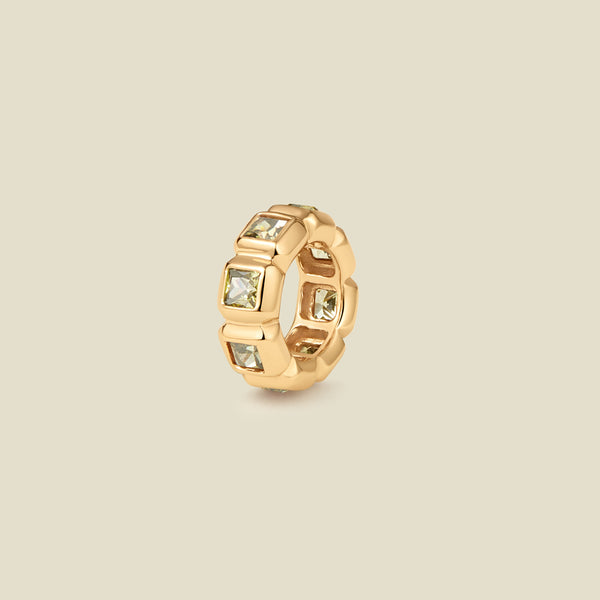 A gold ring charm with multiple bezel set square-cut light green gemstones set side by side to create the band, shown on a solid beige background.
