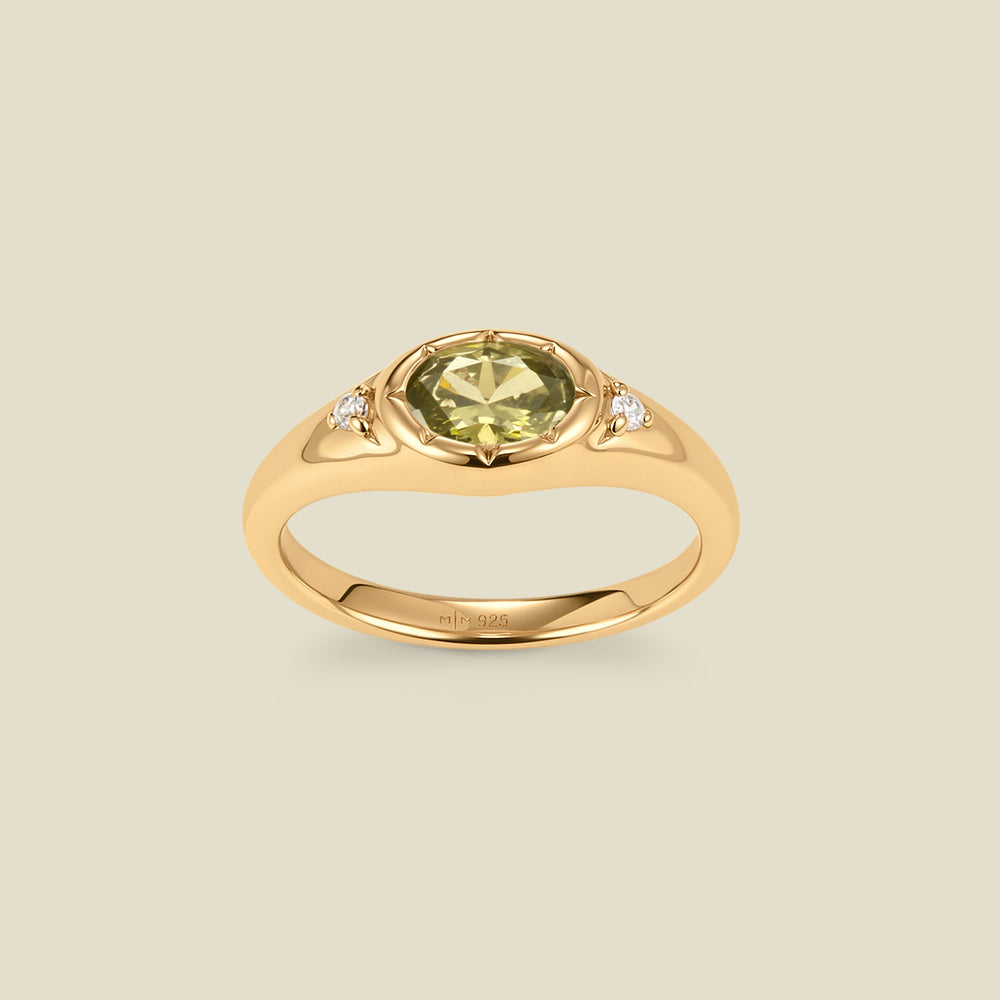 A gold signet ring with a light green center oval bezel-set gemstone, and two small clear accent stones on either side, set against a solid beige background.