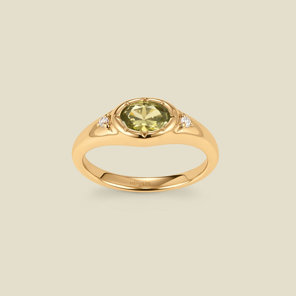 A gold signet ring with a light green center oval bezel-set gemstone, and two small clear accent stones on either side, set against a solid beige background.