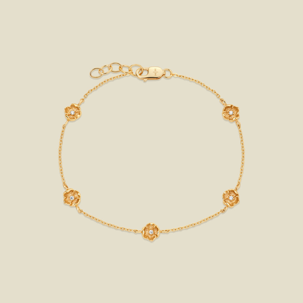 A delicate gold chain bracelet featuring five ornate, textured gold poppy charms with small clear stones, arranged in a circle on a light beige background.
