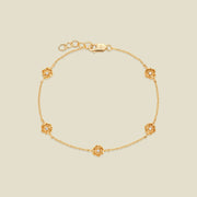 A delicate gold chain bracelet featuring five ornate, textured gold poppy charms with small clear stones, arranged in a circle on a light beige background.