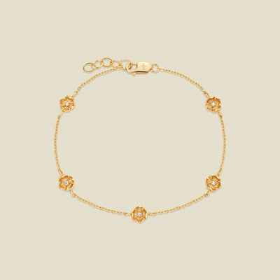 A delicate gold chain bracelet featuring five ornate, textured gold poppy charms with small clear stones, arranged in a circle on a light beige background.